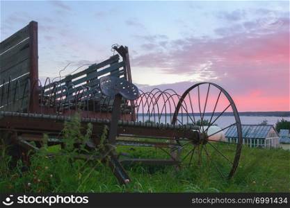 Vintage rusty hay rake with an old wooden wagon on the background of a dramatic pink cloudy sky over the river at sunrise. Ancient agriculture equipment in the countryside. Selective focus.