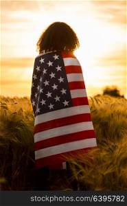 Vintage style photograph of mixed race African American girl teenager female young woman in a field of wheat or barley crops wrapped in USA stars and stripes flag in golden sunset evening sunshine