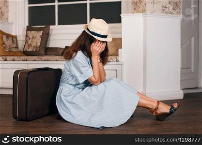 Vintage travelling concept with a young woman hiding her face in hands and sitting on the floor nearby her luggage in the living room. Couch and window on the background.