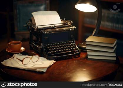Vintage typewriter on wooden table in home office, nobody. Writer workplace in retro style, cup of coffee and glasses, lamp light. Vintage typewriter on wooden table in home office