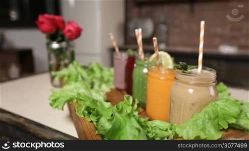 Vintage wooden tray decorated with green salad and different freshly blended vegetable and fruit smoothies in mason jars. Smoothies with striped straws and decor on the table over blurry red roses bouquet in water-glass and rustic kitchen background.