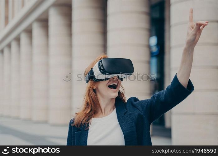 Virtual reality in business. Amazed redhead businesswoman with opened mouth using VR glasses outside, trying to touch something and smiling, blurred buildings on background. Amazed redhead businesswoman with opened mouth using VR glasses outside