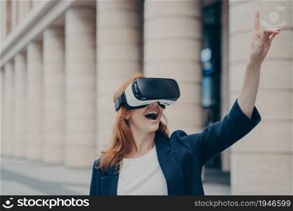 Virtual reality in business. Amazed redhead businesswoman with opened mouth using VR glasses outside, trying to touch something and smiling, blurred buildings on background. Amazed redhead businesswoman with opened mouth using VR glasses outside
