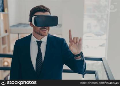 Virtual reality in business. Smiling successful businessman in suit standing in office interior and using VR glasses for work, trying to touch something in air, getting new cyberspace experience. Smiling successful businessman in suit standing in office interior and using VR glasses for work