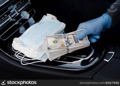 Virus concept. Hand in rubber gloves holds stack of dollar banknotes, earns money as sells medical masks, poses in car. Covid epidemic. Speculation at market of necessary products. Quarantine period