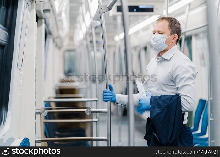 Virus hygiene and safety concept. Serious man dressed in elegant outfit, disposable mask and rubber gloves, touches handrail in underground carriage, commutes to office during quarantine time