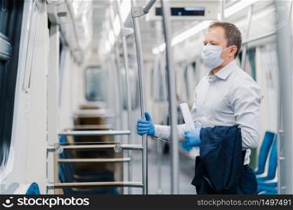 Virus hygiene and safety concept. Serious man dressed in elegant outfit, disposable mask and rubber gloves, touches handrail in underground carriage, commutes to office during quarantine time