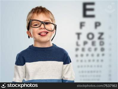 vision and children concept - portrait of smiling little boy with crookedly worn glasses showing tongue over eye test chart background. little boy in glasses showing tongue