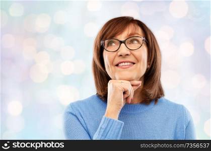 vision and old people concept - portrait of smiling senior woman in glasses looking up and dreaming over festive lights background. portrait of senior woman in glasses dreaming