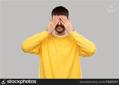 vision and people concept - man in yellow sweatshirt closing his eyes with hands over grey background. man in yellow sweatshirt closing his eyes by hands