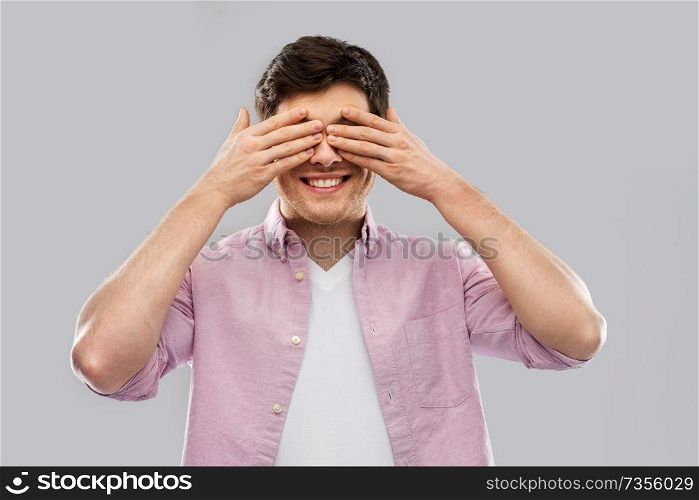 vision and people concept - smiling man closing his eyes by hands over grey background. man closing his eyes by hands over grey background