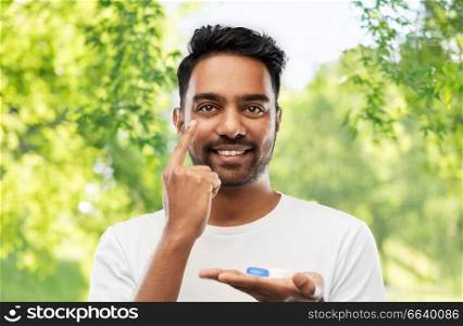 vision, eyesight, ophthalmology and people concept - smiling young indian man applying contact lenses over green natural background. young indian man applying contact lenses