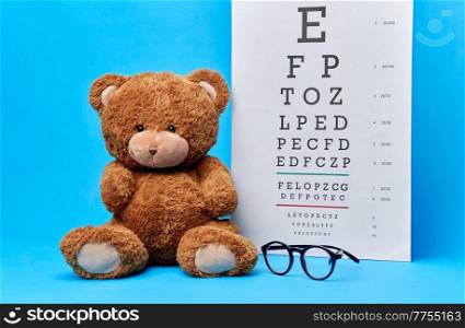 vision, health and childhood concept - brown teddy bear with glasses and eye chart over blue background. teddy bear toy with glasses and eye chart on blue