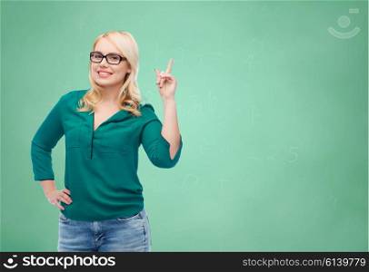 vision, optics, education, gesture and people concept - smiling young woman with eyeglasses pointing finger up over green school chalk board background