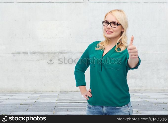 vision, optics, education, gesture and people concept - smiling young woman with eyeglasses showing thumbs up over gray concrete wall background