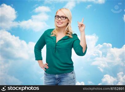 vision, optics, education, gesture and people concept - smiling young woman with eyeglasses pointing finger up over blue sky and clouds background