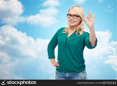 vision, optics, education, gesture and people concept - smiling young woman with eyeglasses showing ok over blue sky and clouds background
