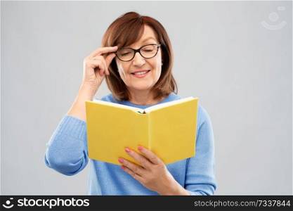 vision, wisdom and old people concept - portrait of smiling senior woman in glasses reading book over grey background. portrait of senior woman in glasses reading book