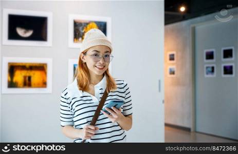 Visitor woman standing takes picture art gallery collection in front framed paintings pictures with mobile phone, Asian female watching at photo frame with smartphone at artwork gallery show