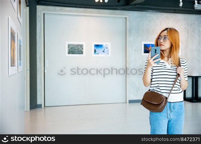 Visitor woman standing takes picture art gallery collection in front framed paintings pictures on white wall with mobile phone, people watch at photo frame with smartphone at artwork gallery show