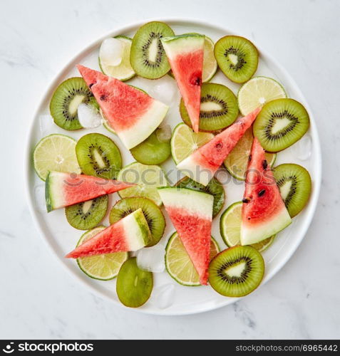 Vitamin organic slices of watermelon, kiwi, lime and ice cubes in a plate on a gray marble background. Ingredients for a healthy summer dessert. Flat lay. Juicy slices of fruit, lime, kiwi, watermelon and ice cubes in a plate on a gray marble background. Flat lay