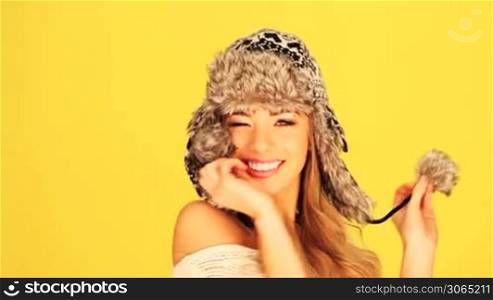 Vivacious laughing blonde woman playing with the pom poms on a woolly winter hat, copyspace on yellow