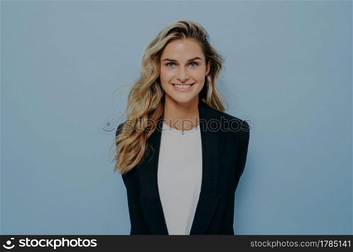 Vivacious young blond woman in black stylish blazer with happy beaming smile standing with hands behind her back like hidding some surprise, isolated over blue studio background. Vivacious blond woman in stylish blazer with happy beaming smile standing with hands behind her back