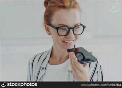 Voice search. Close up shot of redhead woman in transparent glasses speaks to virtual assistant holds mobile phone near mouth poses against white background smiles broadly. Technology communication. Redhead woman in transparent glasses speaks to virtual assistant holds mobile phone near mouth