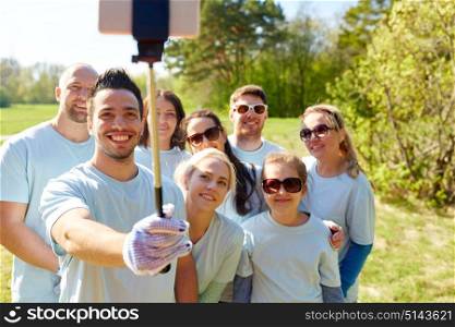 volunteering, charity and people concept - group of happy volunteers taking picture by smartphone and selfie stick in park. group of volunteers taking smartphone selfie
