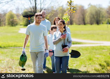 volunteering, charity, people and ecology concept - group of happy volunteers with tree seedlings and rake walking in park
