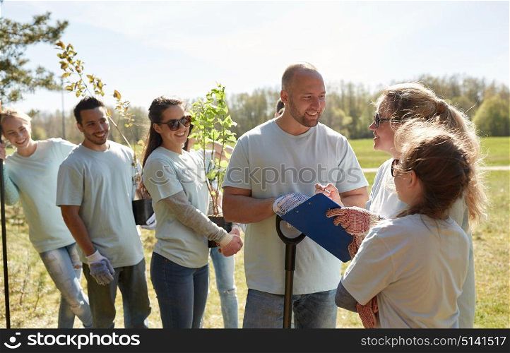 volunteering, charity, people and ecology concept - group of happy volunteers with tree seedlings and clipboard talking in park. group of volunteers with tree seedlings in park