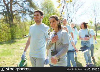 volunteering, charity, people and ecology concept - group of happy volunteers with tree seedlings and shovel walking in park. group of volunteers with trees and rake in park