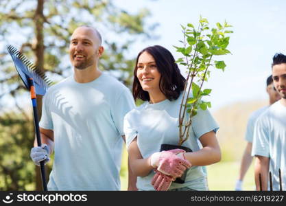 volunteering, charity, people and ecology concept - group of happy volunteers with tree seedlings and rake walking in park