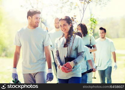 volunteering, charity, people and ecology concept - group of happy volunteers with tree seedlings and rake walking in park. group of volunteers with trees and rake in park