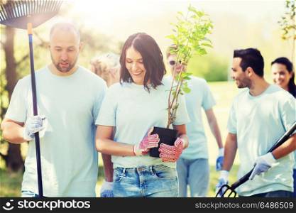 volunteering, charity, people and ecology concept - group of happy volunteers with tree seedlings and rake walking in park. group of volunteers with trees and rake in park. group of volunteers with trees and rake in park