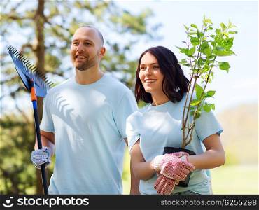 volunteering, charity, people and ecology concept - happy couple volunteers with tree seedlings and rake walking in park