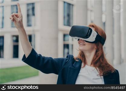 VR technology for your business. Happy young businesswoman using virtual reality goggles while standing outdoors, managing project with finger, testing headset, office buildings in blurred background. Happy young businesswoman using virtual reality goggles while standing outdoors