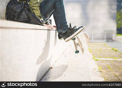 Waist down view of young male urban skate boarder sitting on wall flipping skateboard with feet