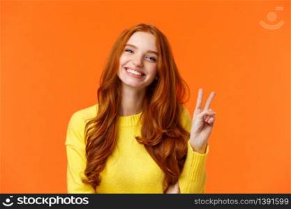 Waist-up cheerful gorgeous redhead woman with long curly red hair, showing peace sign and smiling happily, express positivity, like winter holidays, standing orange background.. Waist-up cheerful gorgeous redhead woman with long curly red hair, showing peace sign and smiling happily, express positivity, like winter holidays, standing orange background