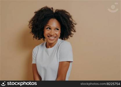 Waist up of cute curly african woman with toothy smile dressed in white t shirt looking aside with shyness and expressing positive emotions, isolated over beige background. Face expressions concept. Cute african woman with toothy smile looking aside with shyness, isolated over beige background