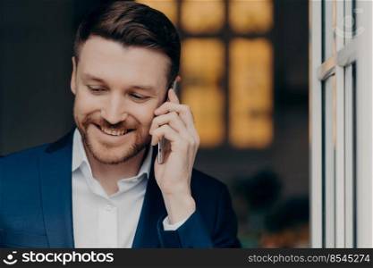 Waist up of smiling bearded business person in suit having corporate mobile conversation, enjoying good talk while standing indoors and looking down with happy expression. Business concept. Happy stylish businessman talking on mobile phone with pleasure