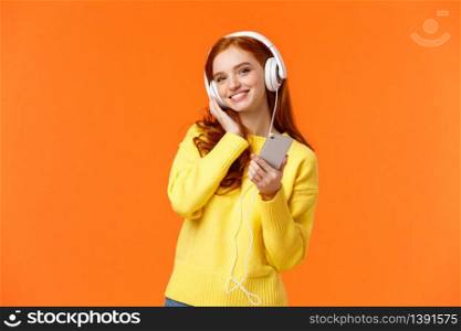 Waist-up portrait cheerful lovely young woman with red hair listen music in headphones, tilt head and smiling satisfied, holding smartphone, pick song for tender calm evening, orange background.. Waist-up portrait cheerful lovely young woman with red hair listen music in headphones, tilt head and smiling satisfied, holding smartphone, pick song for tender calm evening, orange background
