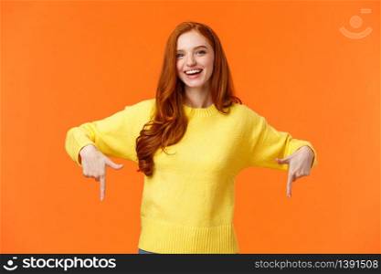 Waist-up portrait cheerful redhead female student in yellow sweater, inviting check-out event, recommend proudct, chrismas sale, holiday promo, pointing down and smiling, orange background.. Waist-up portrait cheerful redhead female student in yellow sweater, inviting check-out event, recommend proudct, chrismas sale, holiday promo, pointing down and smiling, orange background