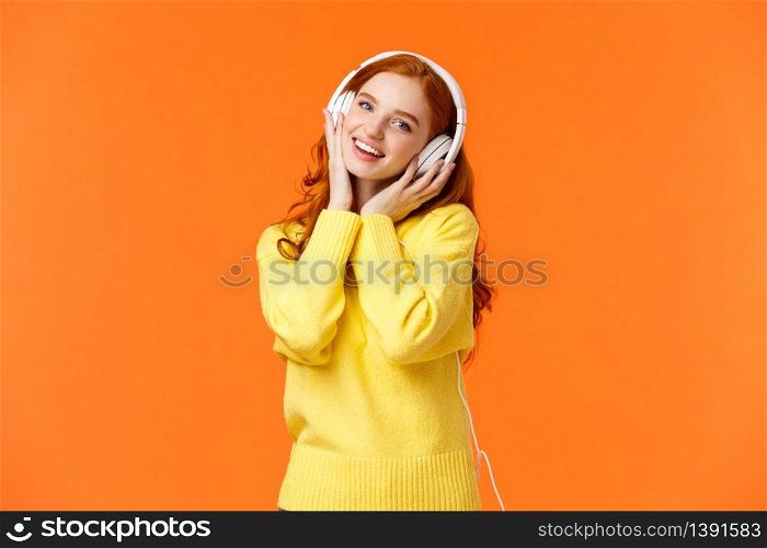 Waist-up portrait cute teenage redhead girl in white headphones, tilt head listen music, touch earphones and smiling camera, singing along favorite song, standing orange background. Copy space. Waist-up portrait cute teenage redhead girl in white headphones, tilt head listen music, touch earphones and smiling camera, singing along favorite song, standing orange background