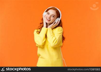 Waist-up portrait cute teenage redhead girl in white headphones, tilt head listen music, touch earphones and smiling camera, singing along favorite song, standing orange background. Copy space. Waist-up portrait cute teenage redhead girl in white headphones, tilt head listen music, touch earphones and smiling camera, singing along favorite song, standing orange background