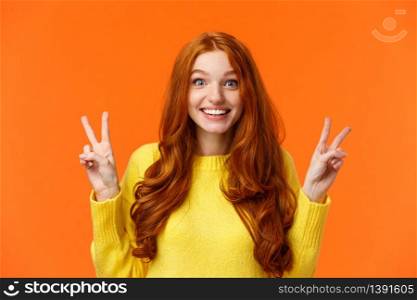 Waist-up portrait lovely and cheerful pretty redhead woman showing peace gestures, smiling camera, travel abroad during winter holidays posing near sightseeing, orange background.. Waist-up portrait lovely and cheerful pretty redhead woman showing peace gestures, smiling camera, travel abroad during winter holidays posing near sightseeing, orange background