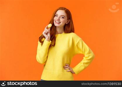 Waist-up portrait lovely redhead girl holding tasty yellow macaron near mouth and smiling joyfully, like eating sweets, buy dessert for romantic valentines date, posing cheerful orange background.. Waist-up portrait lovely redhead girl holding tasty yellow macaron near mouth and smiling joyfully, like eating sweets, buy dessert for romantic valentines date, posing cheerful orange background