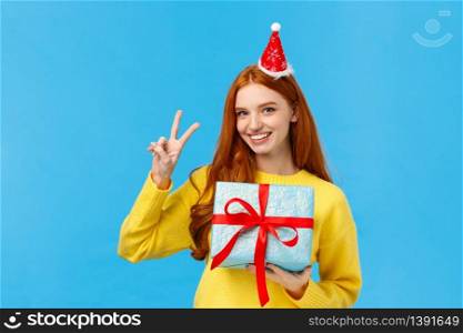 Waist-up portrait of cute happy redhead woman receive gift in wrapped box, celebrating christmas holidays, wearing fancy new year hat and showing peace sign, smiling joyfully, blue background.. Waist-up portrait of cute happy redhead woman receive gift in wrapped box, celebrating christmas holidays, wearing fancy new year hat and showing peace sign, smiling joyfully, blue background