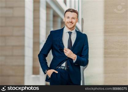 Waist up portrait of successful male lawyer comes on meeting with client, wears elegant formal clothes, holds shades, looks happily at camera, keeps hand in pocket. Business and people concept