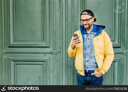 Waist up portrait of trendy hipster with beard dressed in casual clothes holding hand in pocket looking through news on his web-enabled smartphone, chatting with friends using his smartphone